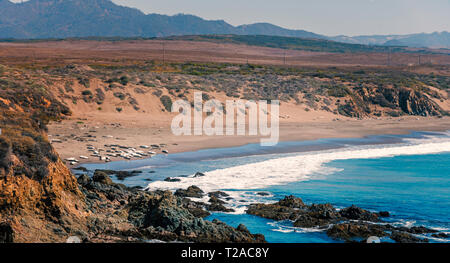 Overlooking blue ocean with waves breaking onto rocks and beach below, fields with hazy mountains beyond under bright sky. Stock Photo