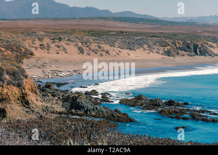 Overlooking blue ocean waves rolling onto rocky shore with hazy mountains beyond under hazy blue sky. Stock Photo
