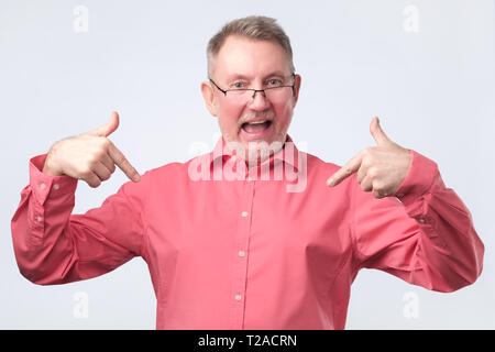 Senior european man in red shirt and glasses is proud of himself. Husband pointing on himself with index finger. Stock Photo