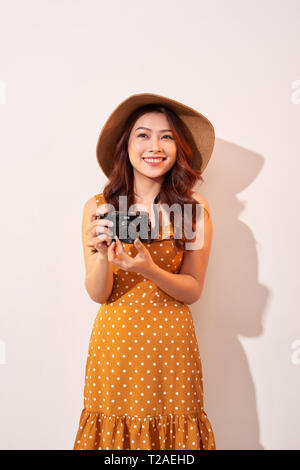 Portrait of cheerful smiling young woman taking photo with inspiration and wearing summer dress. Girl holding retro camera. Model posing on beige back Stock Photo