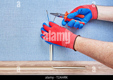 Worker in protective gloves is cutting ends of copper wiring in cable channel of floor plinth with working tool. Stock Photo