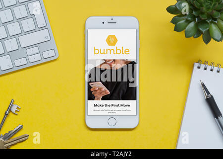 An iPhone showing the Bumble website rests on a yellow background table with a keyboard, keys, notepad and plant (Editorial use only). Stock Photo