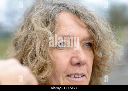 Close up portrait of smiling middle aged woman with curly hair, wrinkled face and blue eyes, standing outdoors and looking to the side Stock Photo