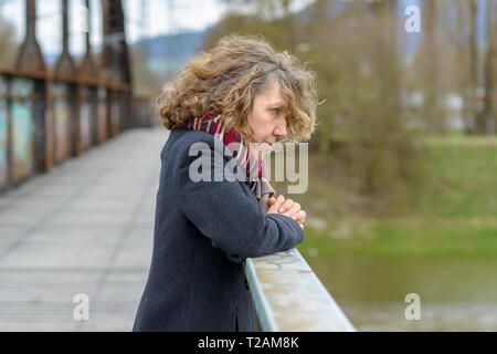 Introspective woman standing thinking deeply as she leans on the railing of a pedestrian bridge gazing over the edge in a warm coat and scarf Stock Photo