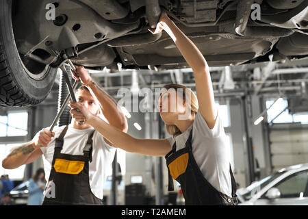 Professional mechanics standing under automobile and working in workshop. Young female worker with strong male colleague checking chassis of car in au Stock Photo