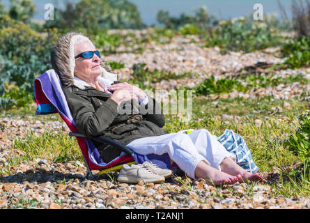 Senior woman wearing a coat sitting on a beach chair on a cold day in Spring in the UK. Stock Photo