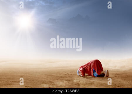 Rear view of asian muslim man praying in prostration position on desert with sun rays and dark sky background Stock Photo