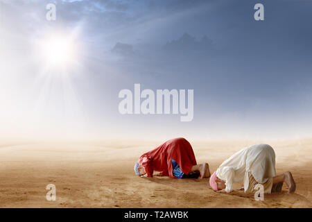 Rear view of two asian muslim man praying in prostration position on desert with sun rays and dark sky background Stock Photo