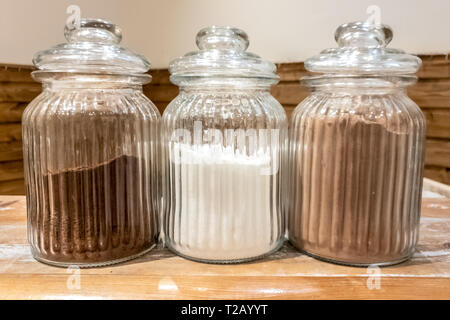 Three half full transparent glass jars with cafe, sugar and cacao with lids on a wooden table in the cafeteria Stock Photo