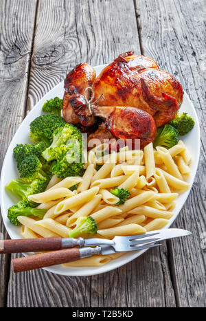 a Whole roasted chicken with broccoli and penne rigate on a white platter with fork and knife, on a grey rustic wooden table, vertical view from above Stock Photo