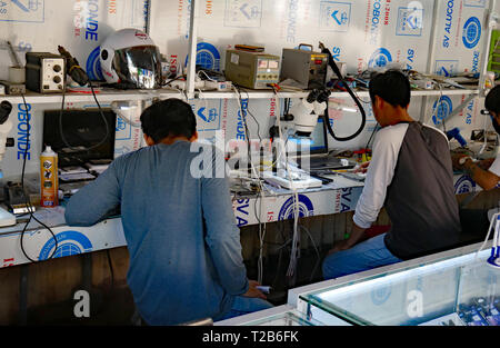 Young technicians working at a cluttered workbench repairing mobile phones. Battambang, Cambodia 15-12-2018 Stock Photo