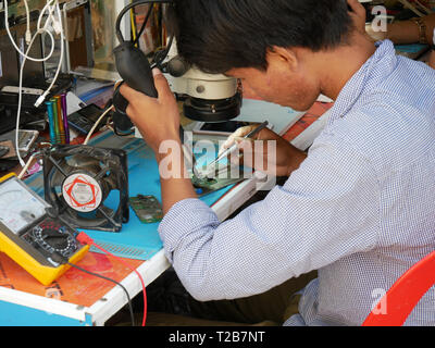 A Khmer technician repairing a mobile phone at the roadside in an open fronted shop in a Cambodian town. Battambang, Cambodia 13-12-2018 Stock Photo