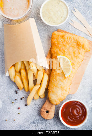Traditional British Fish and Chips with tartar sauce abd glass of craft lager beer and tomato ketchup on chopping board on white stone table backgroun Stock Photo
