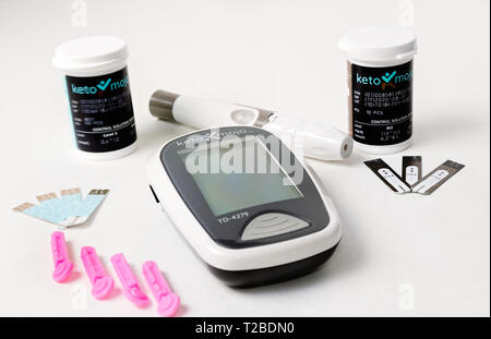 A Keto-Mojo ketone and blood glucose meter is pictured on white, along with ketone and glucose test strips, disposable needles, and lancet device. Stock Photo
