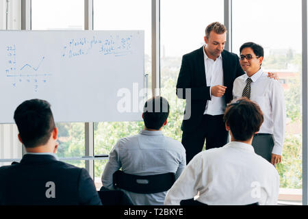Caucasian business manager introduces new Asian businessman in front of seminar meeting with many employees listening in company office setting Stock Photo
