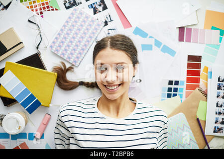 Above view portrait of contemporary young woman smiling at camera while lying on top of art and design supplies, copy space Stock Photo