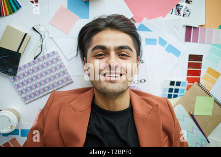 Top view portrait of contemporary Middle-Eastern man looking at camera while lying over  art and design supplies, copy space Stock Photo