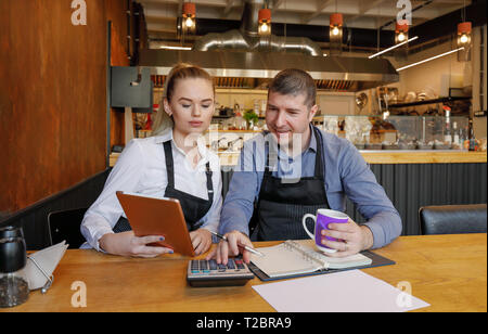 Young woman and her husband sitting at table with tablet and crumpled papers doing accounts for their small restaurant late in evening. Restaurant own Stock Photo