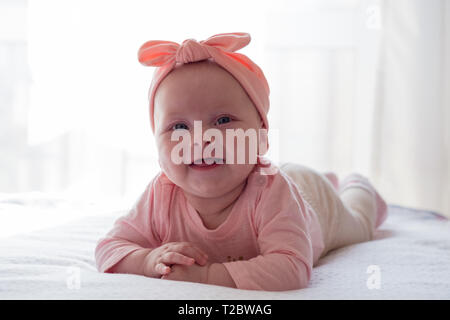 A newborn girl with a pink bandage on her head is lying on her stomach. The child smiles. Stock Photo