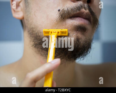 closeup man use yellow shaver shaving messy beard and mustache on his face in bathroom Stock Photo
