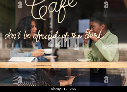 Two diverse girlfriends enjoying  coffee and talking together while sitting at a counter by the window of a dark trendy cafe Stock Photo