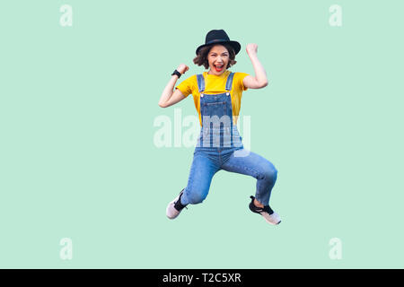 Portrait of screaming happy suprised young girl in blue denim overalls, yellow shirt and black hat jumping, looking and celebraiting her victory. indo Stock Photo