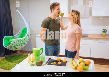 Young blonde woman gives her husband an organic apple while cooking in the kitchen. Healthy eating family. Vegetarian meal. Diet detox Stock Photo