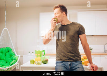 Young smiling handsome man bites organic apple while cooking fresh fruits in the kitchen. Healthy eating. Vegetarian meal. Diet detox Stock Photo