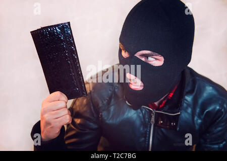 Young ill-intended man wearing black mask holds the black leather women purse and looks to his loot. Robber has stolen the wallet. Half shot of a thie Stock Photo
