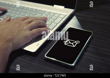 SMS password for network access on phone while typing on laptop. Generic icon of mail on black-screened smartphone lying on the office table close to  Stock Photo