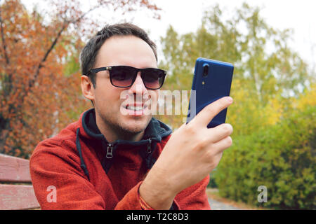 Hipster man using mobile phone in autumn park, send a fun message. Student chatting with his collegues from university. male student chatting with fri Stock Photo