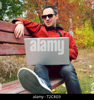 Joyful guy communicates through the Internet on the street. Programmer enjoy with notebook sitting in autumn park. Handsome man working on his laptop. Stock Photo