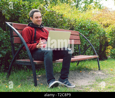 Joyful guy communicates through the Internet on the street. Programmer enjoy with notebook sitting in autumn park. Handsome man working on his laptop. Stock Photo