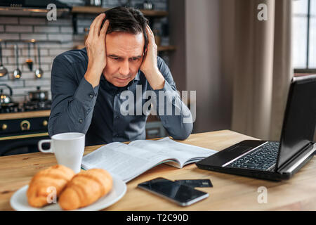 Serious concentrated man sit at table in kitchen. He hold hands on hears and read journal. Man work Stock Photo