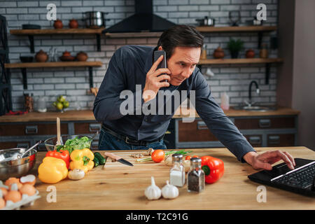 Adult businessman stand at kitchen table desk and reach laptop keyboard. He talk on phone. Guy is concentrated. Colorful vegetables lying on desk Stock Photo