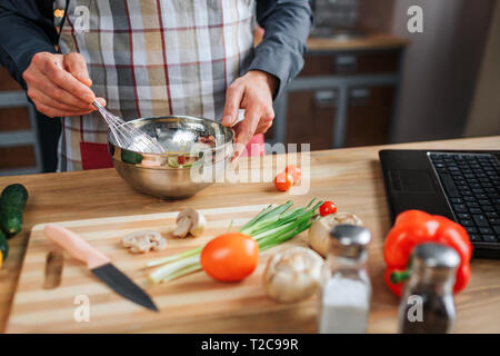 Close up of man's hands blending eggs in bowl. He work at table in kitchen. Guy wear apron. Colorful vegetables and laptop of keyboard on desk Stock Photo
