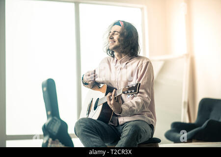 Cheerful handsome young guitarist sitting in bright room Stock Photo