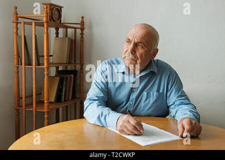 Old man writing notes or deciding whether to sign a paper document on the table in front of him as he looks to the side with a thoughtful engrossed ex Stock Photo