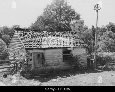 Black and white photograph of a small wooden shack, with a shingled roof, brick chimney stack, and a bell set on top of a tall pole at right; likely a former slave quarters that was converted for use as a dairy storage facility known as a milk house or depot; located in Bardstown, Kentucky, USA; photographed by Marion Post Wolcott, under the sponsorship of the United States' Farm Security Administration, August, 1940. From the New York Public Library. () Stock Photo