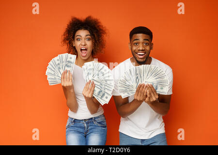 Big win! Shocked and happy black guy and girl holding lots of money and shouting, orange background Stock Photo