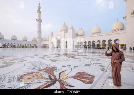 Young woman taking pictures at the Sheikh Zayed Grand Mosque inner courtyard with ornate flower themed floor mosaics, Abu Dhabi, United Arab Emirates Stock Photo