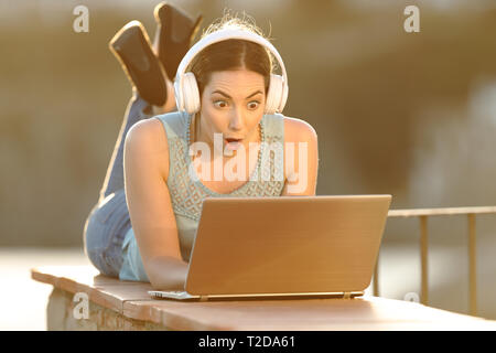 Surprised girl with headphones checking laptop lying on a balcony wall Stock Photo