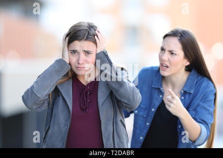 Angry girl scolding her sad friend who looks at camera in the street Stock Photo