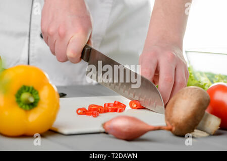 Cook's hands cutting a red chili pepper into slices with a santoku knife. Cooking a dish with bell pepper, onion, mushrooms and tomato. Stock Photo