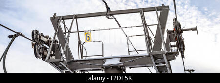 Steel ropes over wheels in mechanism on top of ski chairlift support column, number 6 on yellow plate, bright sky background, wide banner. Stock Photo