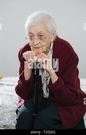 Portrait of a tired senior gray haired woman sitting on bed, leaning chin on a walking stick at home. Resting her head on a cane. Stock Photo