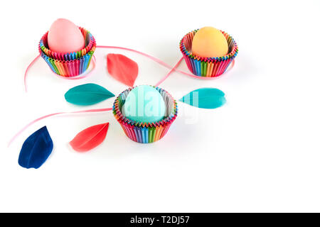 Bright creative Easter decoration of colorful eggs in raibow colored paper forms for cupcakes on white background decorated with feathers. Copy space. Stock Photo