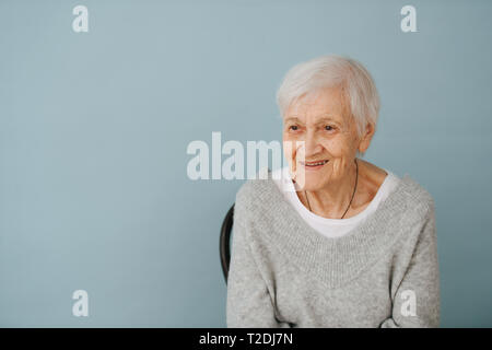 Portrait of smiling elderly grey haired woman seating on a chair, wearing comfortable grey V-type sweater at home. Glasses off. Stock Photo