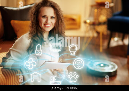 Portrait of smiling young woman with long brunette hair in the modern house using smart home application on tablet PC and robot vacuum cleaning floor  Stock Photo