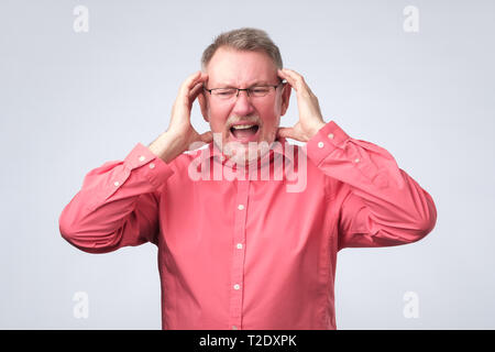 Studio shot of frustrated senior retired man in red shirt and glasses having painful look because of terrible headache, touching head Stock Photo